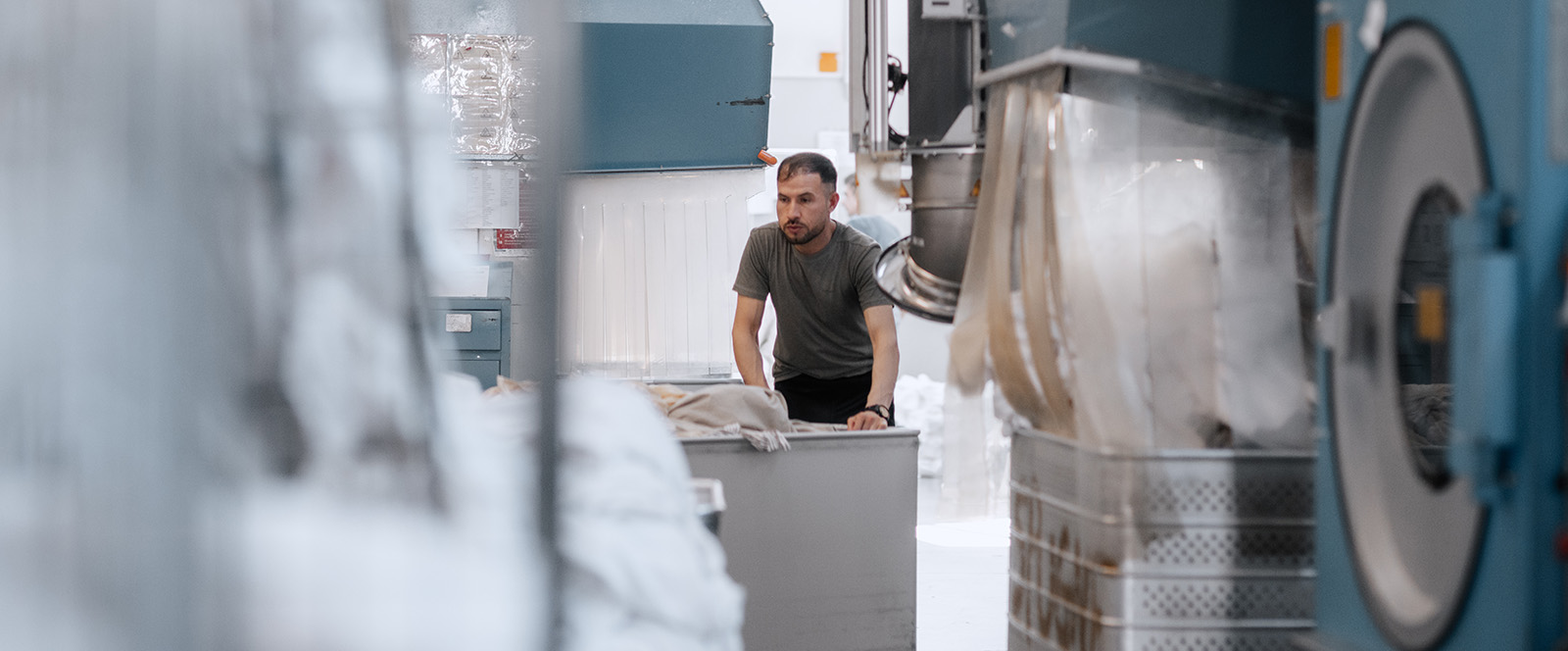 Un uomo spinge un carrello colmo di biancheria, in primo piano una lavatrice industriale.