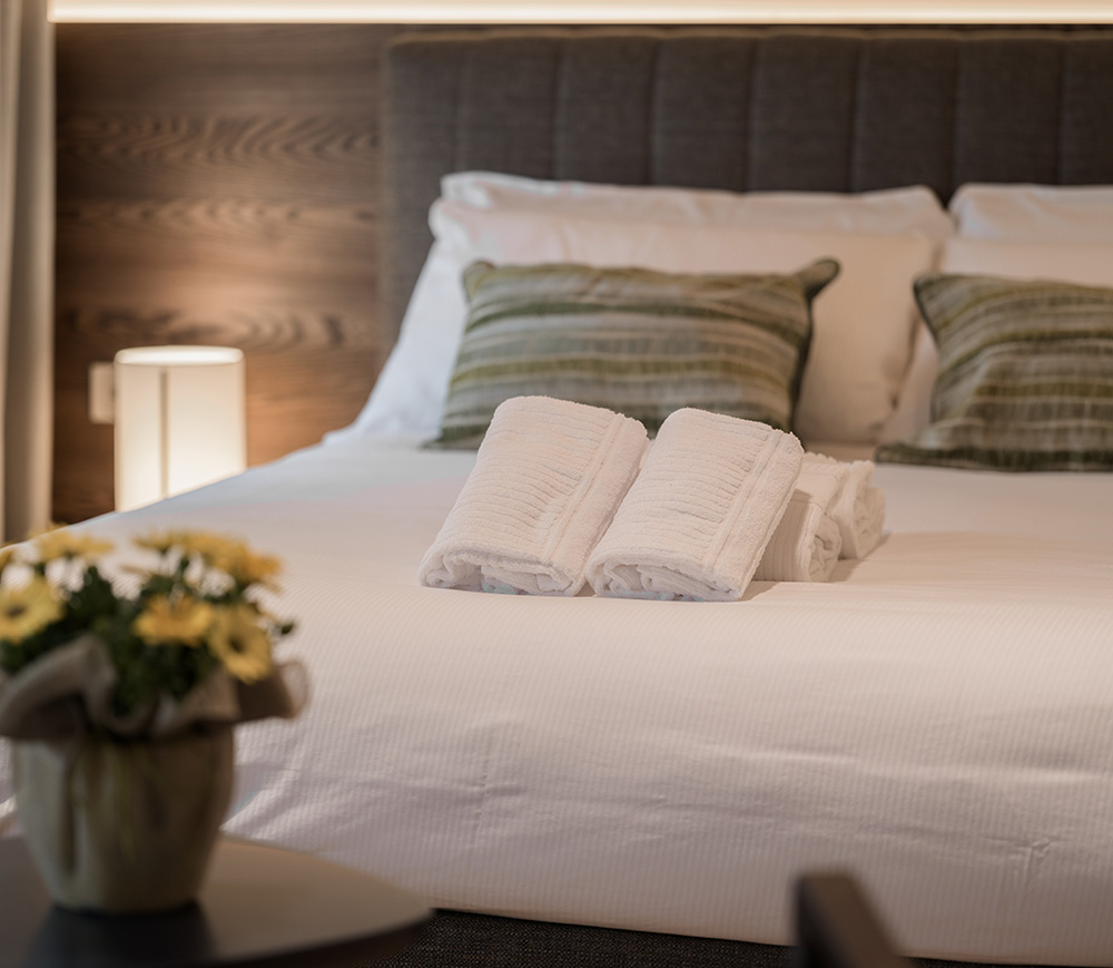 Primo piano di un letto matrimoniale in una camera d'hotel del quale spiccano la biancheria e un set di asciugamani poggiato ai piedi del letto.