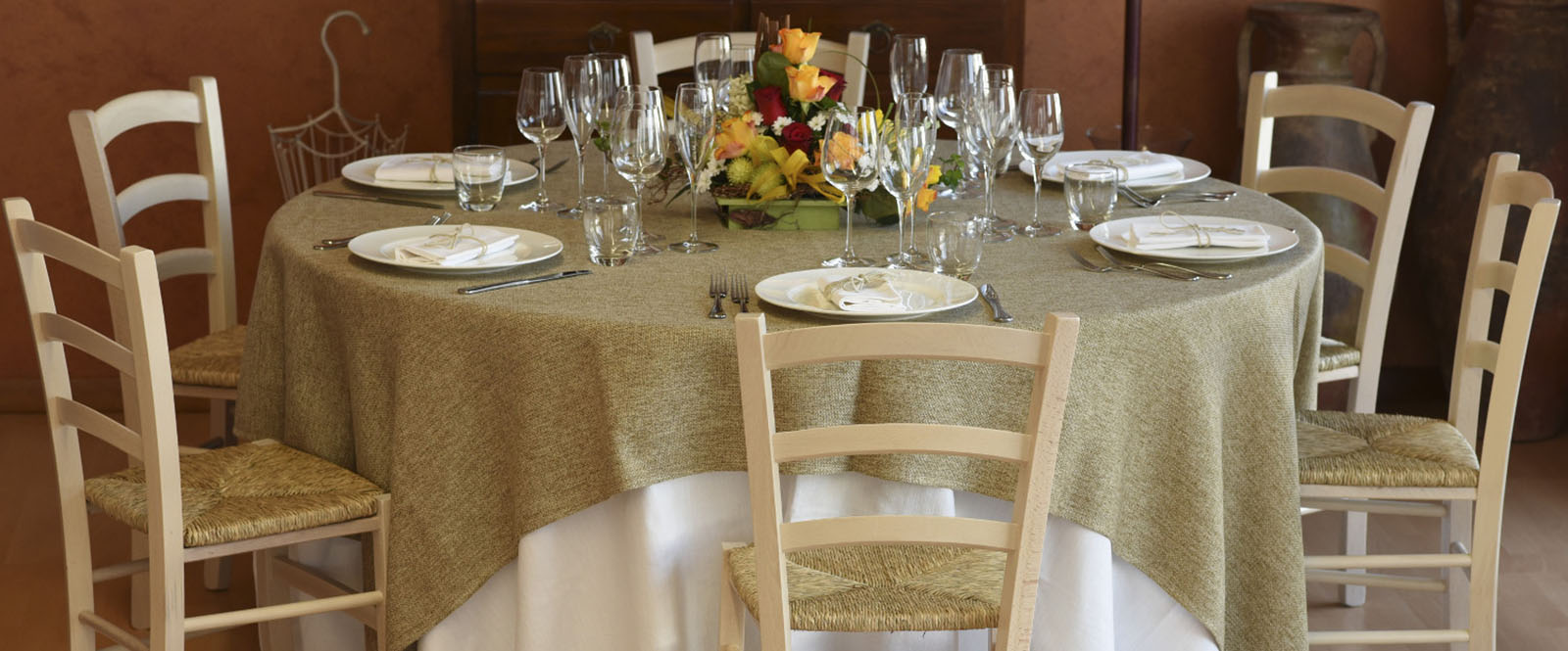 Tavolo apparecchiato per sei persone con una tovaglia in juta, stoviglie, bicchieri e un centro tavola floreale.